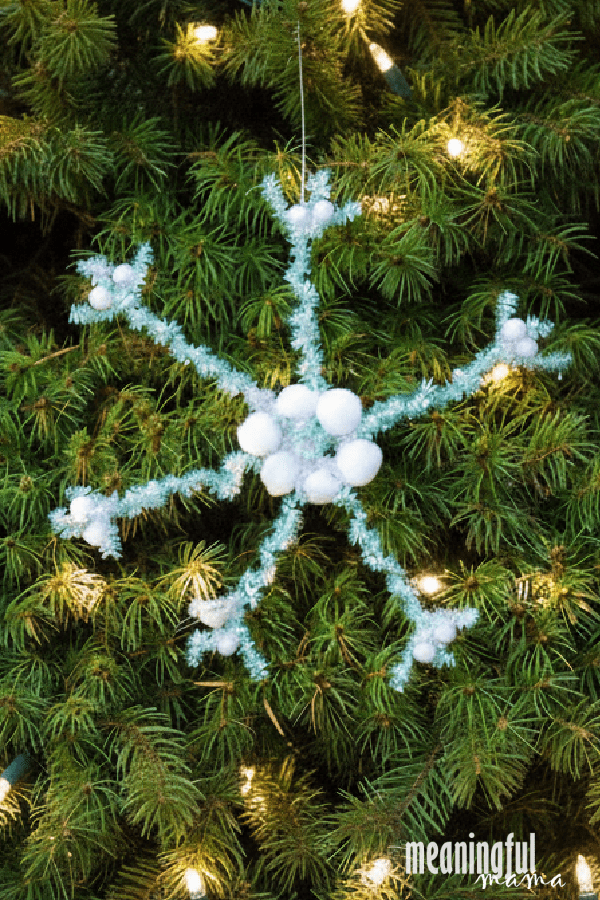 Pipe cleaner snowflake with spiral icy blue arms and white pom poms hanging in a lit Christmas tree with green branches