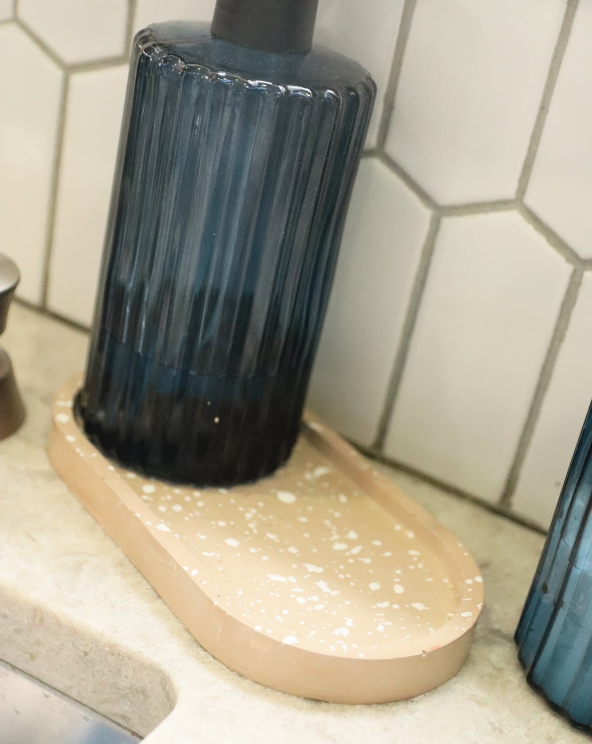Peach speckled plaster tray holding a ribbed blue glass soap pump on a kitchen counter in front of white hexagon tile
