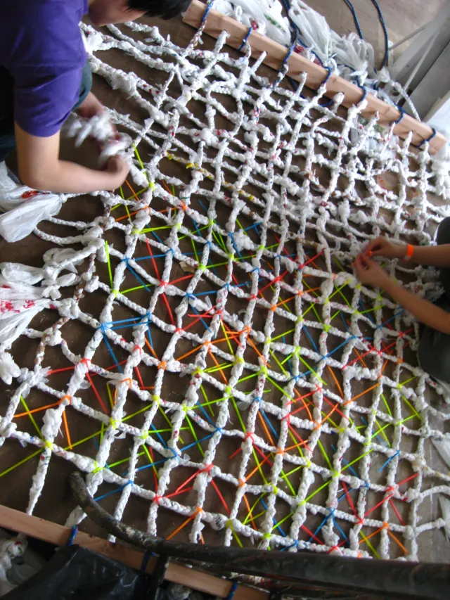 Large diamond net woven from twisted white plastic bag rope with colored ties, two people working on it on the floor