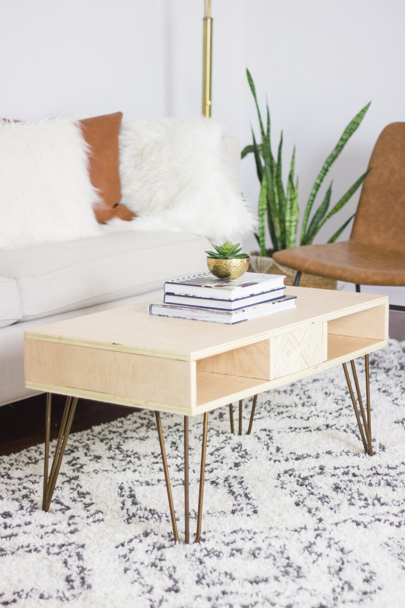 Light plywood coffee table with patterned panel cubbies on gold hairpin legs, topped with books and a plant by a white sofa