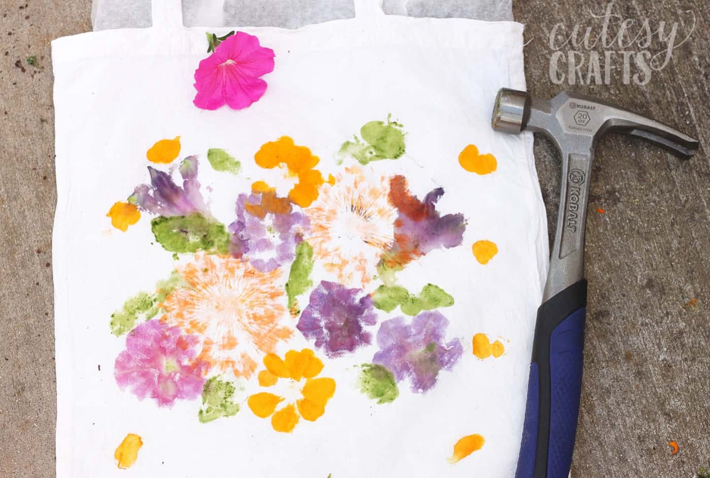Cotton tote bag with pounded flower prints in orange, purple and pink, next to a pink petunia and a claw hammer on concrete