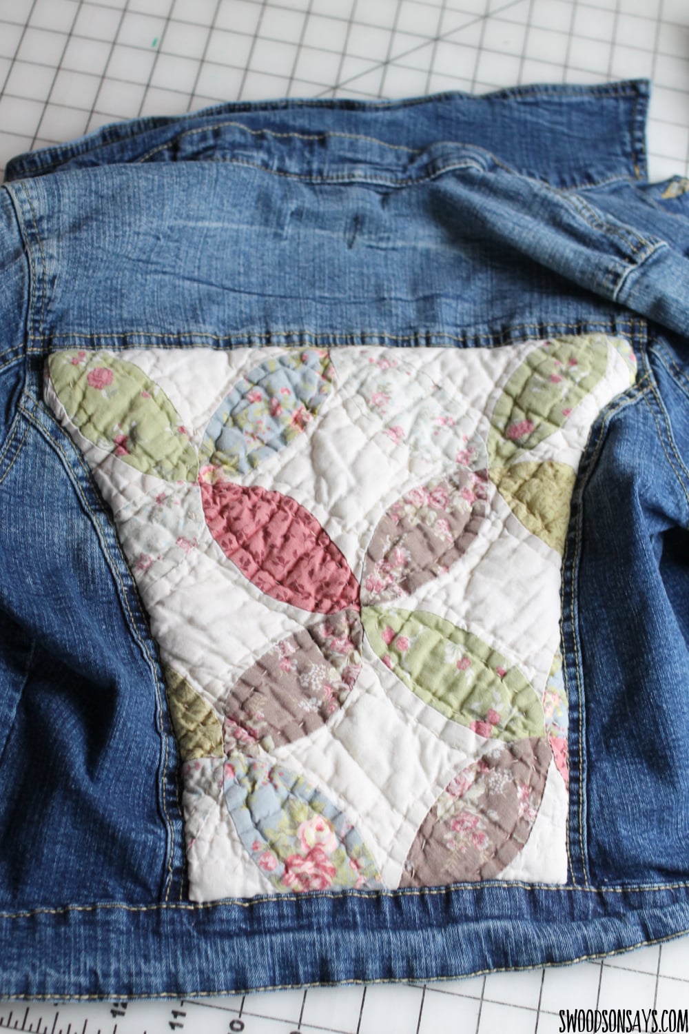 Jean jacket laid flat with a quilted floral patchwork panel in pink, green and cream sewn onto the back on a cutting mat