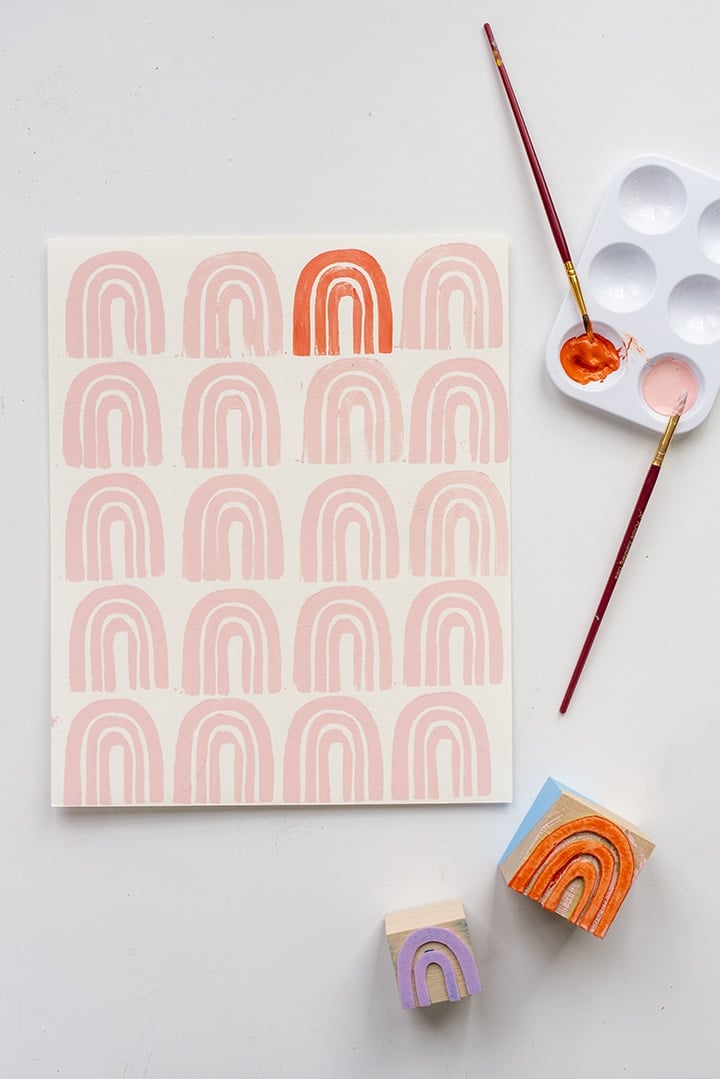 Stamped print of pale pink rainbows with one orange rainbow beside foam rainbow stamps, a paint palette and brushes