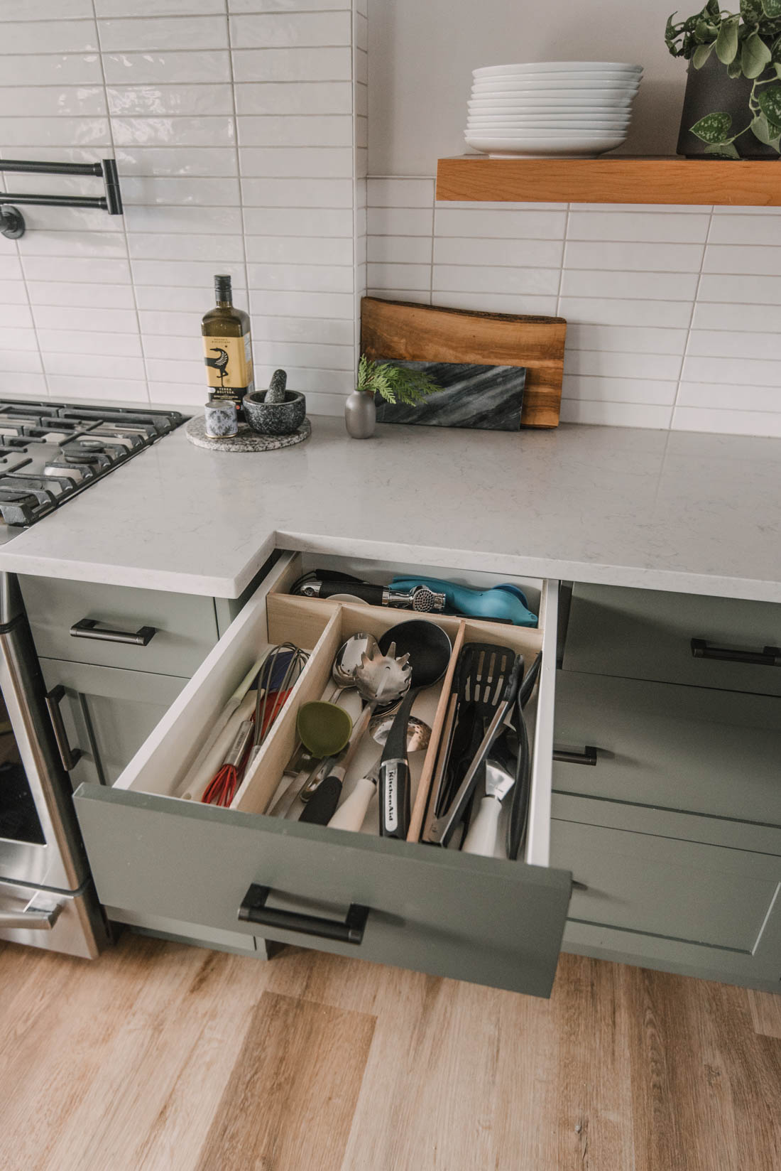 Open sage green kitchen drawer with a removable wood utensil organizer holding spatulas, whisks and tongs