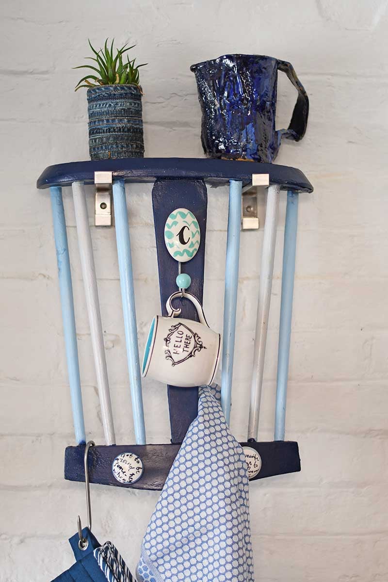 Blue painted chair back hung as a kitchen wall rack with a plant and pitcher on the seat shelf and a mug and towel on hooks