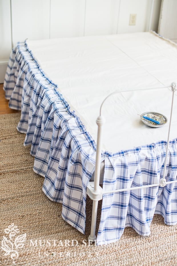 Blue and white plaid ruffled bed skirt on a white iron bed frame over a jute rug, with a pin dish resting on the mattress