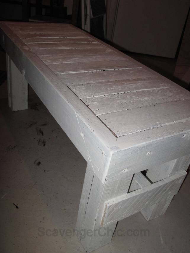 White painted pallet wood bench with chunky stacked legs and a wide plank top, finished and standing on a workshop floor