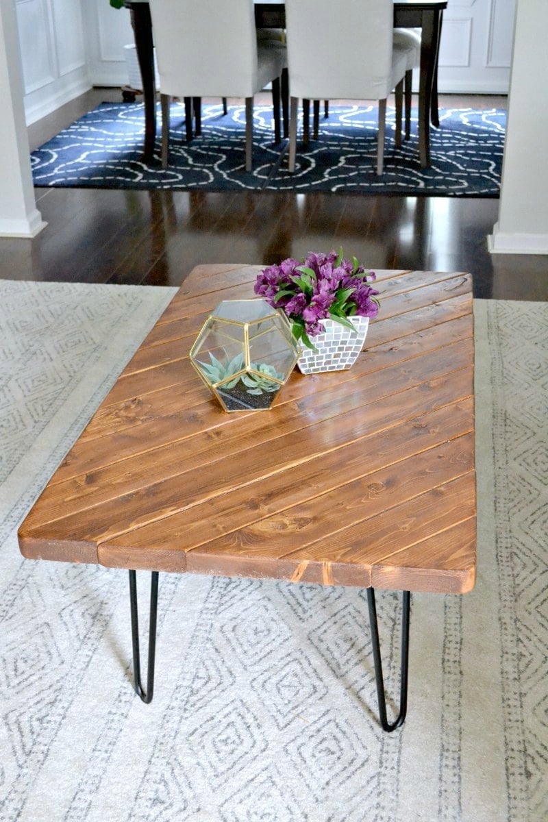 Coffee table with an angled top of stained scrap 2x4 boards on black hairpin legs, topped with purple flowers on a rug