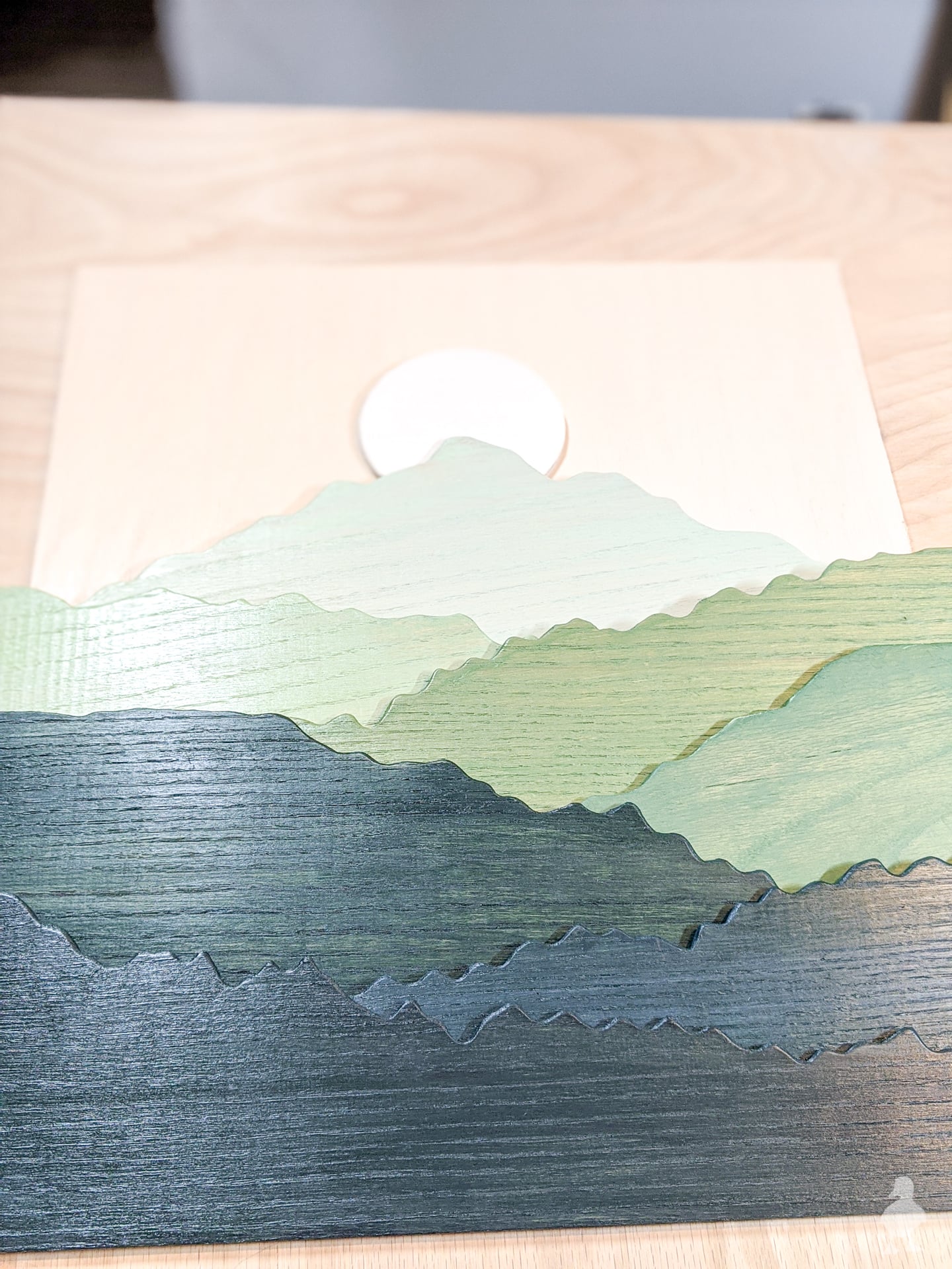 Framed layered wood mountain art in green and navy with a pale sun, leaning against a white wall by a door