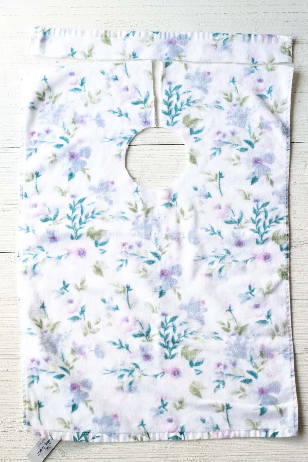 White kitchen towel with lilac and teal floral print sewn into a baby bib with a round neck hole and a slit, on white wood