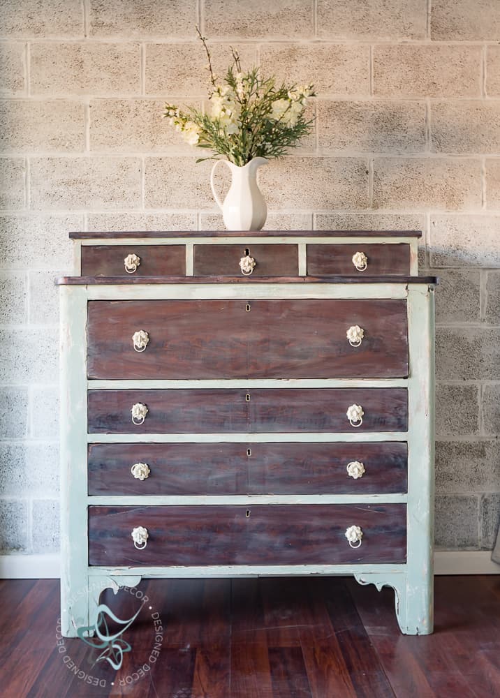Distressed empire dresser with a chippy soft green and cream painted frame, brown washed drawers and a pitcher of flowers