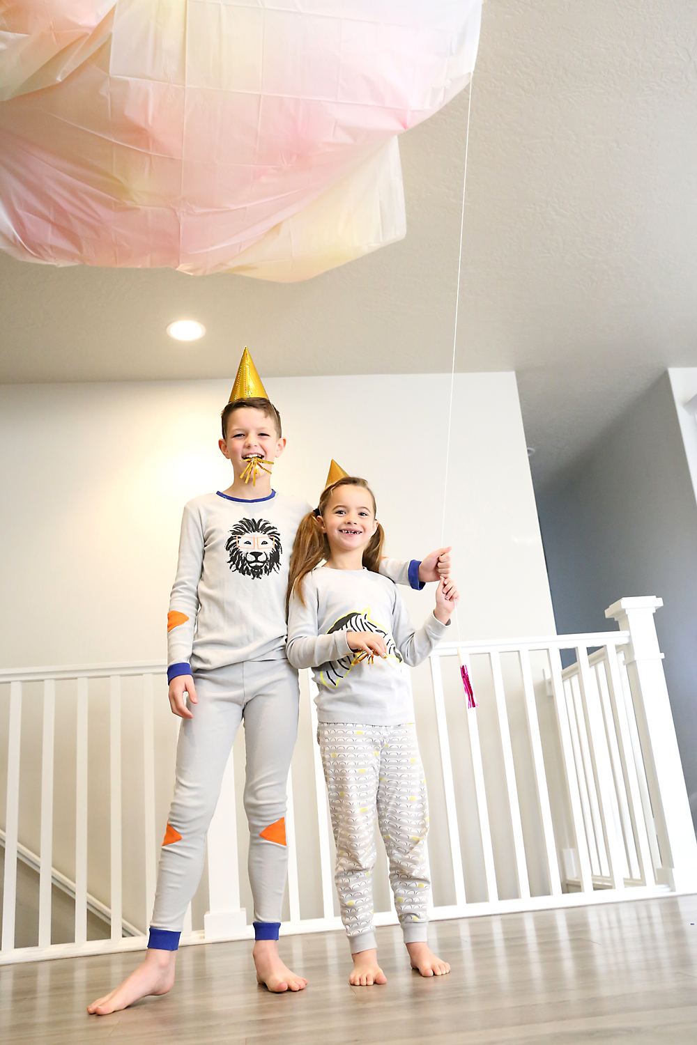Two kids in party hats and pajamas pulling a string to release balloons from a shower curtain taped to the ceiling
