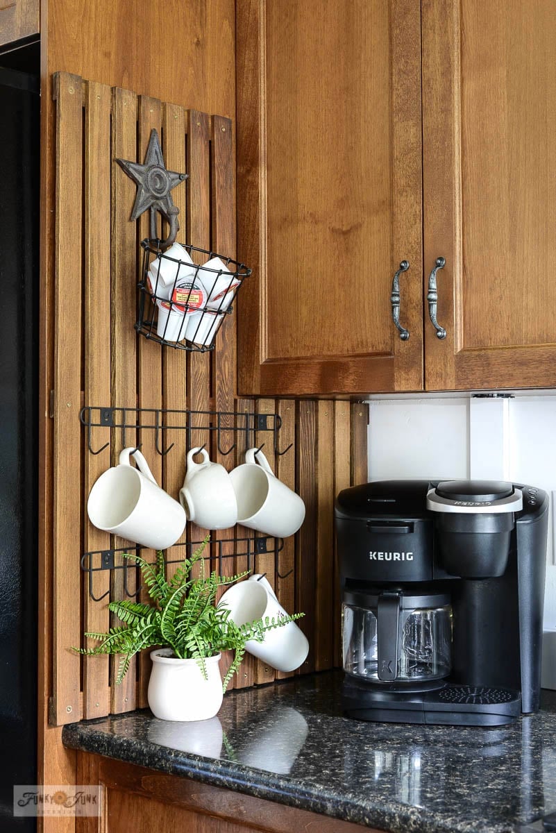 Cedar strip rack on the side of a fridge cabinet with white mugs on black hooks, a wire basket and a coffee maker below