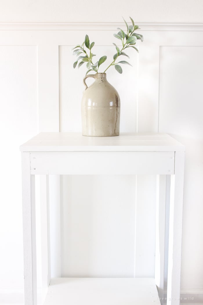 Simple white painted wood side table with a slatted top and a lower shelf, styled with a stoneware jug of olive branches