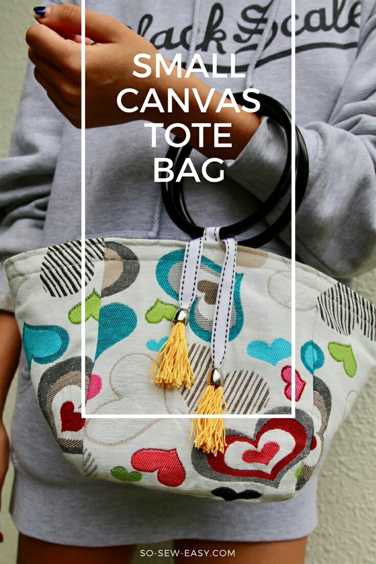 Small heart print canvas tote bag with black round ring handles and two yellow tassels on striped ribbon, held at the waist