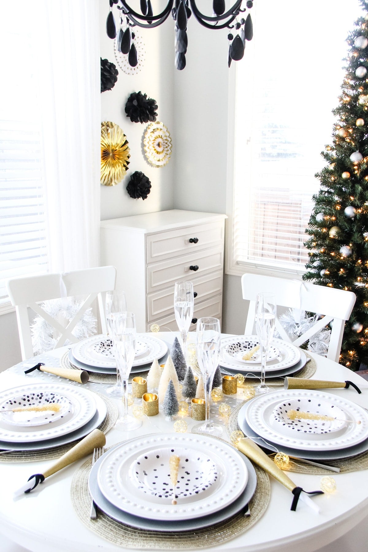 Round white table set in gold, silver and black with bottle brush trees, noisemakers and wreaths on the chairs
