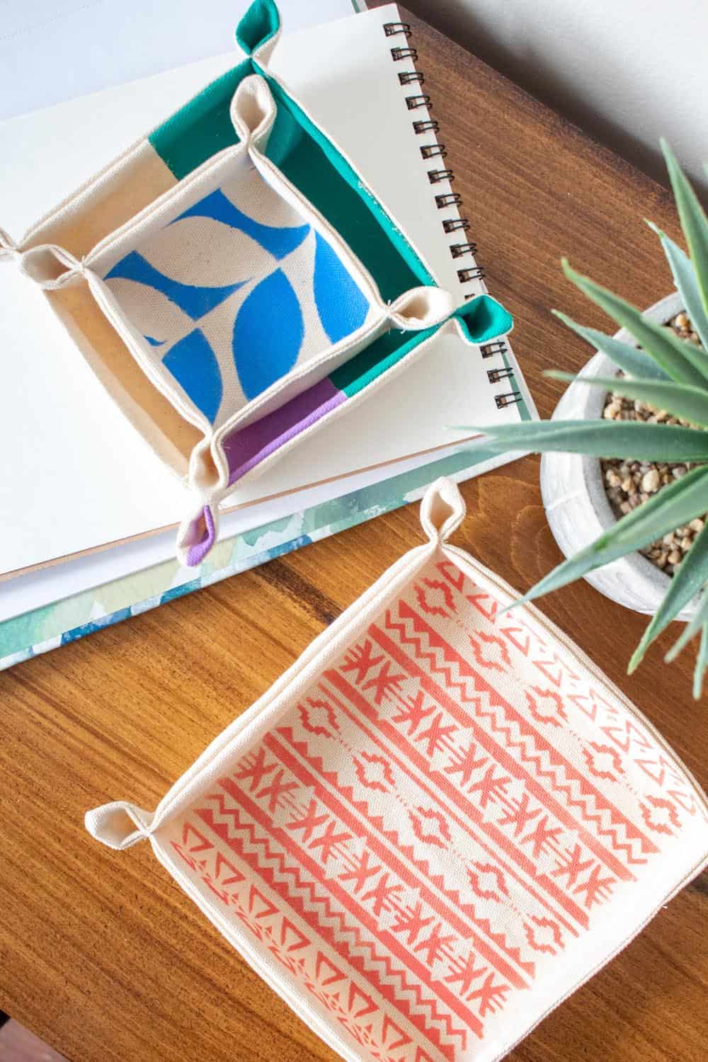 Stenciled Fabric Nesting Trays | Home Sewing Project Stenciled fabric nesting trays with snap corners, one blue and cream, one coral tribal print, on a wood desk beside a plant