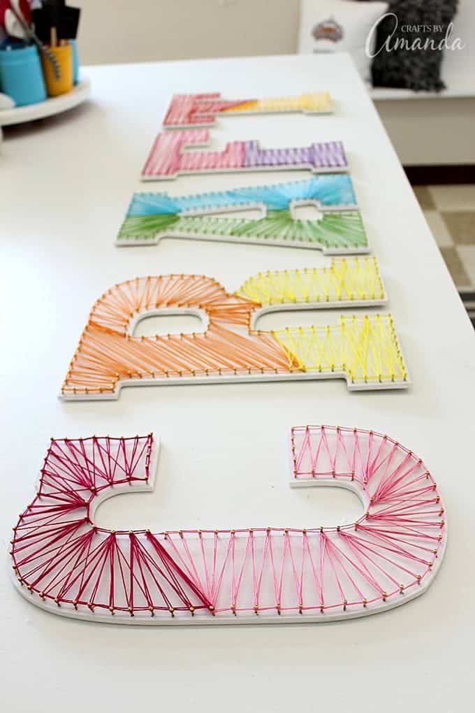 String art letters with rainbow embroidery floss strung between nails, laid in a row on a white craft table