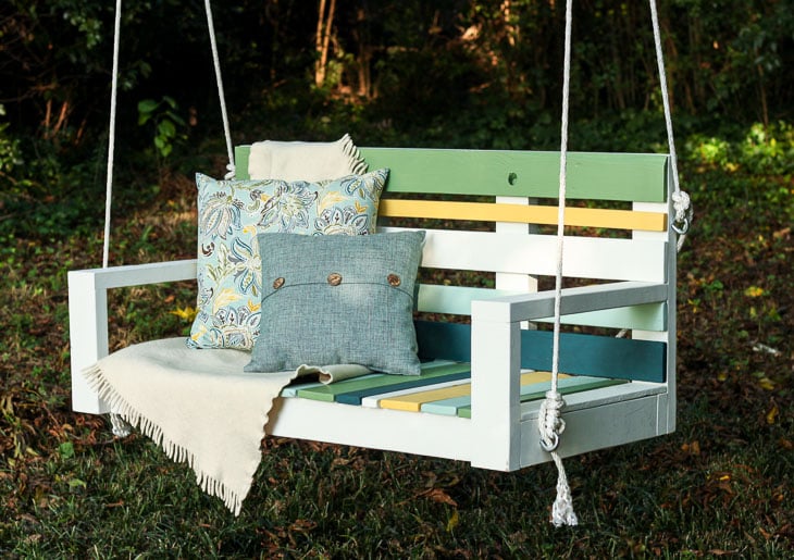 White porch swing with pallet wood slats painted in stripes of green, yellow, blue and teal, hung from a tree with rope
