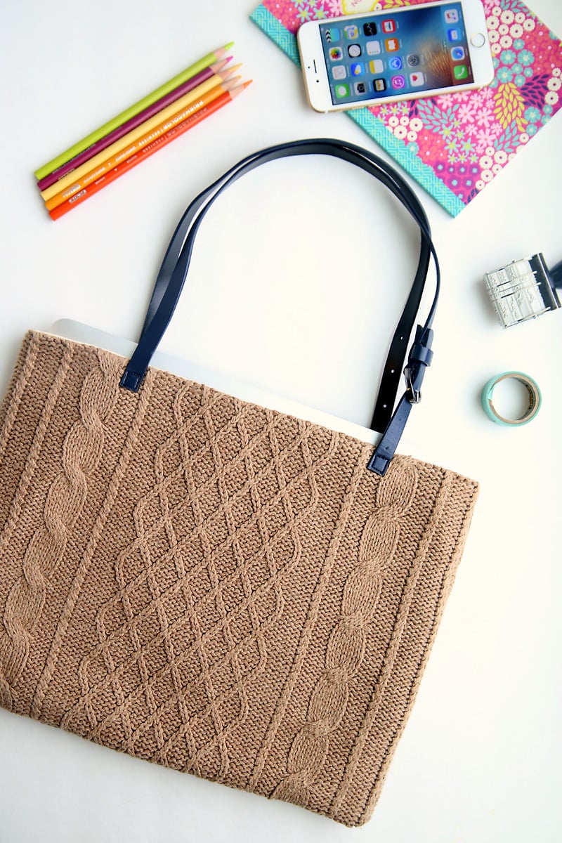 Cable-knit tan sweater tote with navy leather belt handles holding a laptop, a floral notebook and a smartphone