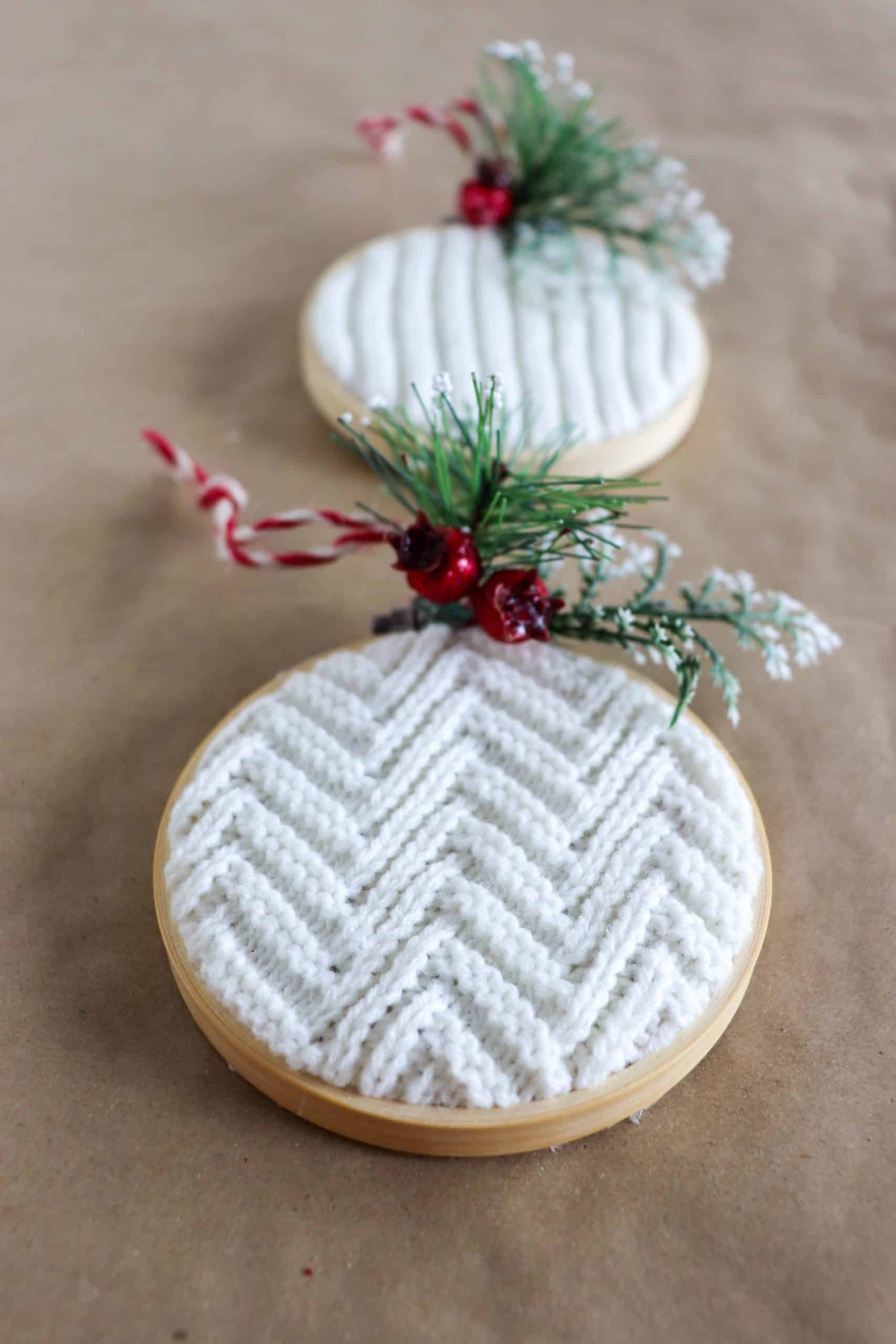 Two embroidery hoop ornaments filled with cream ribbed and chevron sweater knit, topped with faux pine sprigs and red berries