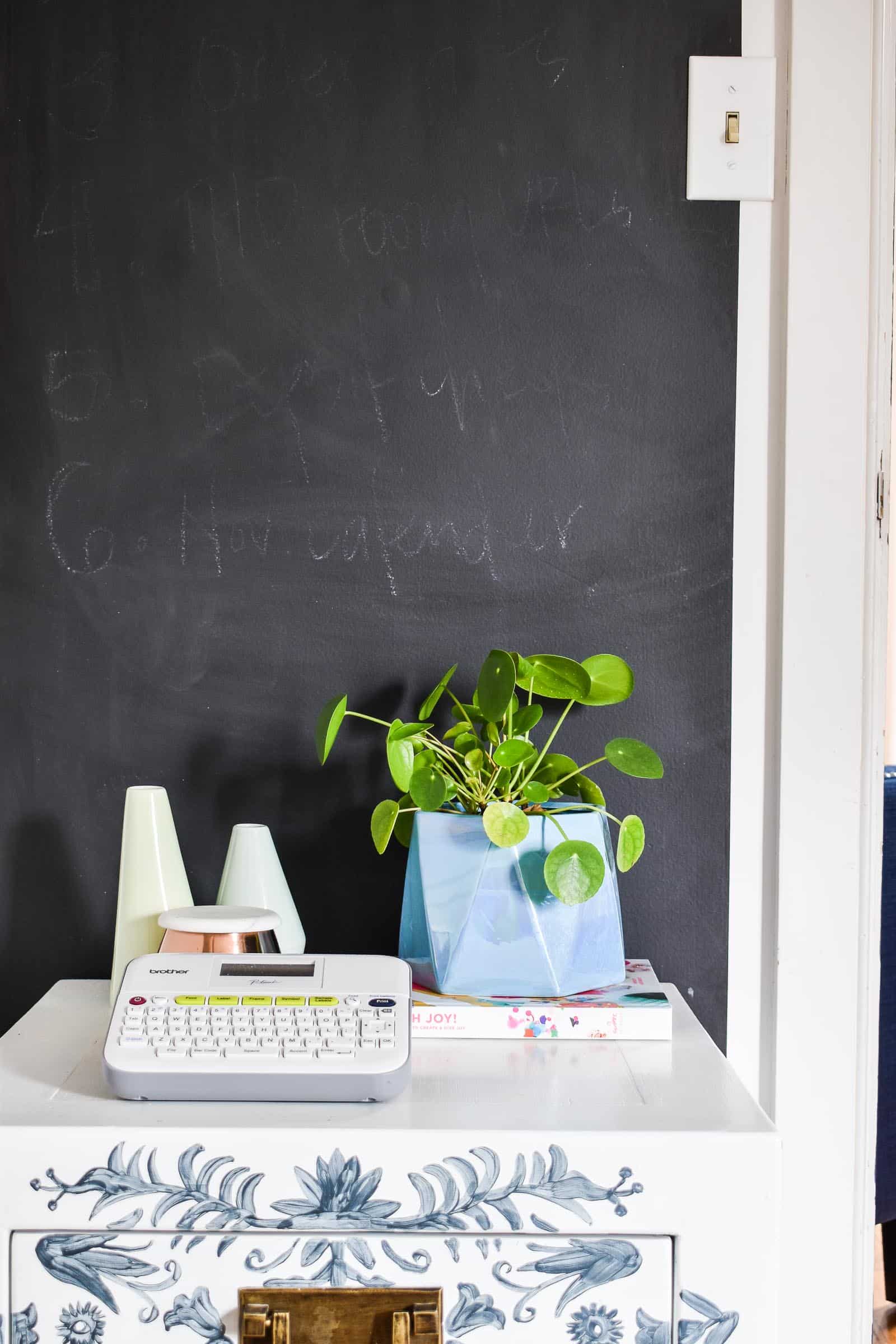 Chalkboard paint wall section in a home office behind a white cabinet with a label maker, a potted plant and white vases