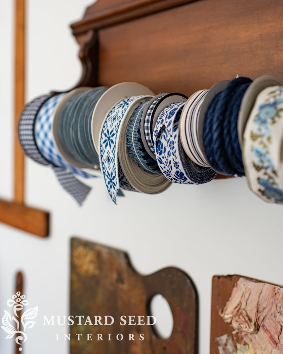 Spools of blue and white patterned ribbon and navy cord lined up on a dark wooden thrifted rack hanging on a white wall