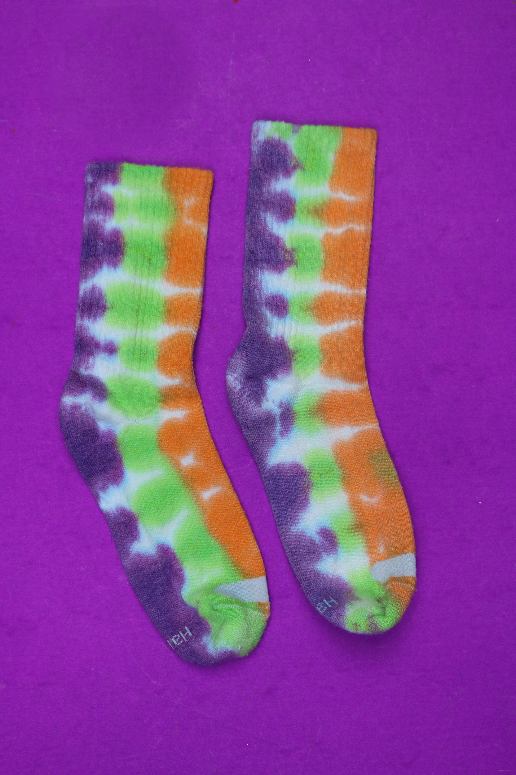 Pair of white crew socks tie-dyed in purple, lime green and orange vertical stripes laid flat on a purple background