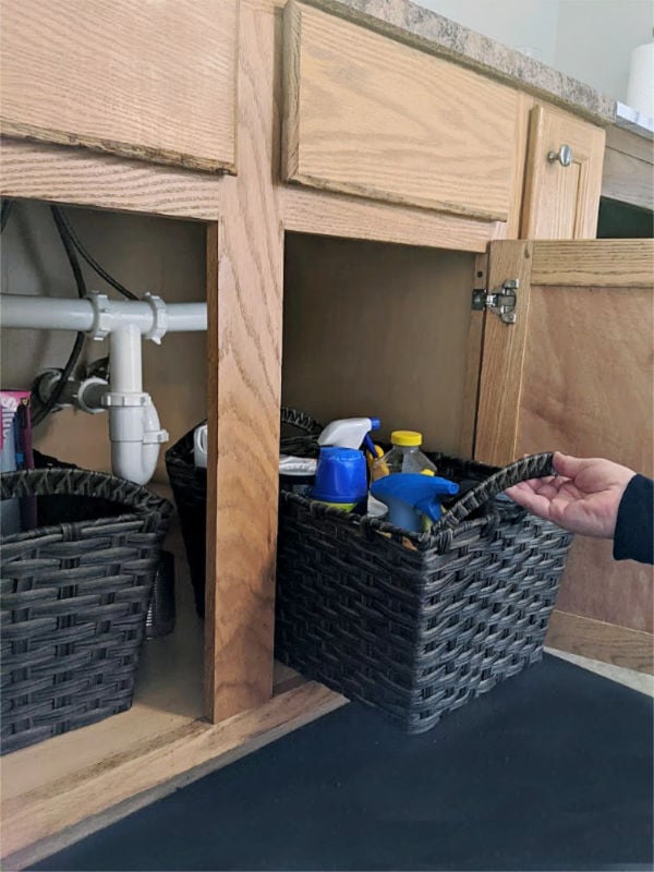 Faux rattan basket pulled out of an open under sink cabinet holding spray bottles and cleaning supplies