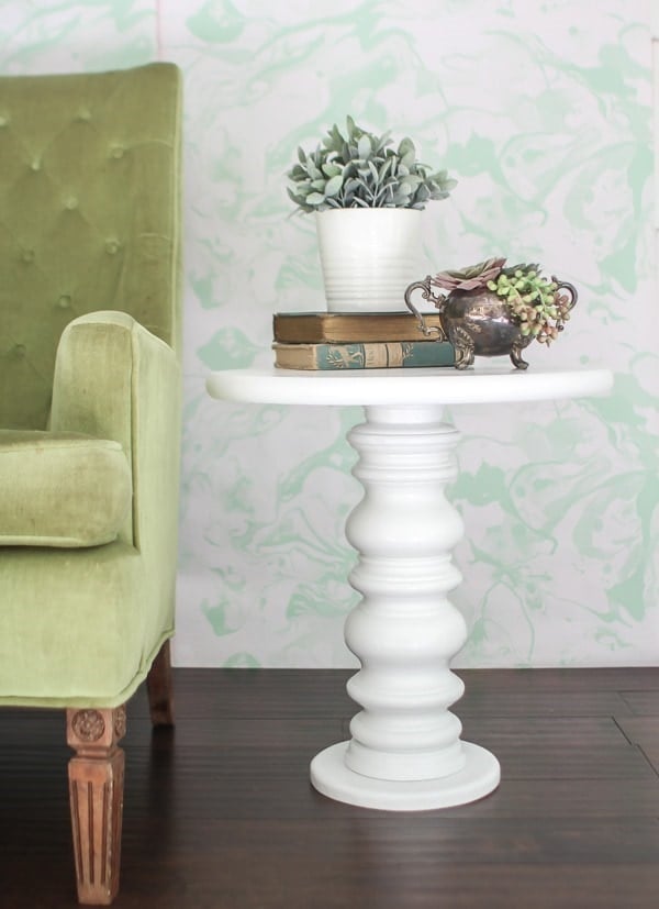 White painted pedestal side table made from a turned wooden lamp base beside a green velvet chair, topped with books