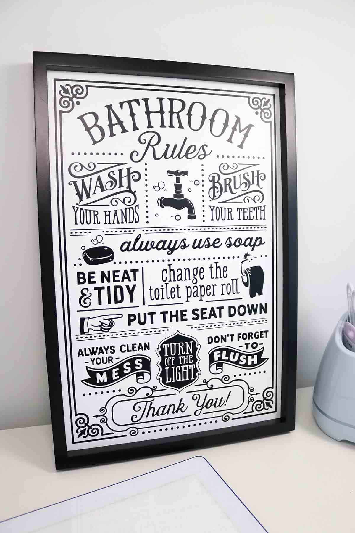 Black framed white bathroom rules sign with vintage lettering and a faucet graphic leaning beside a gray Cricut machine