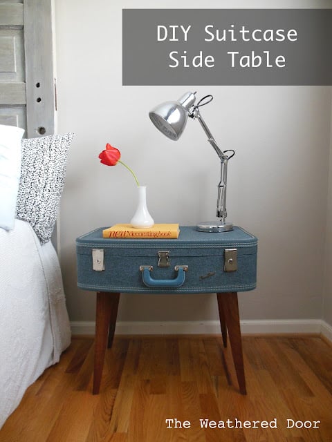 Vintage blue suitcase on four angled wood legs used as a nightstand, topped with a silver desk lamp, a red tulip and a book