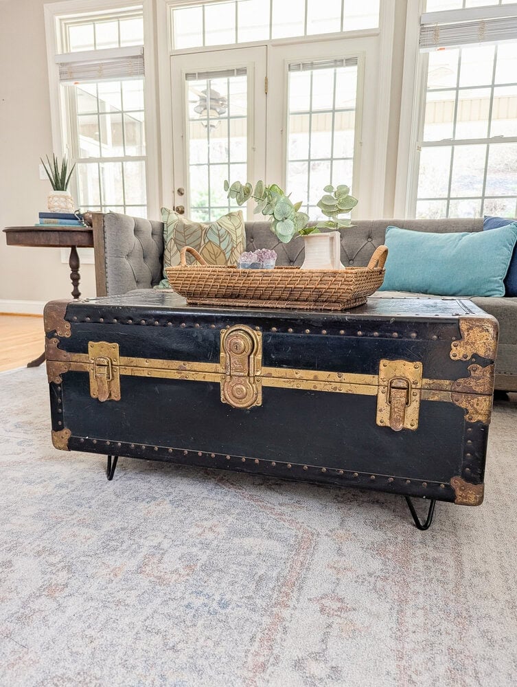 Black vintage trunk with brass fittings on short black hairpin legs, used as a coffee table with a woven tray by a sofa