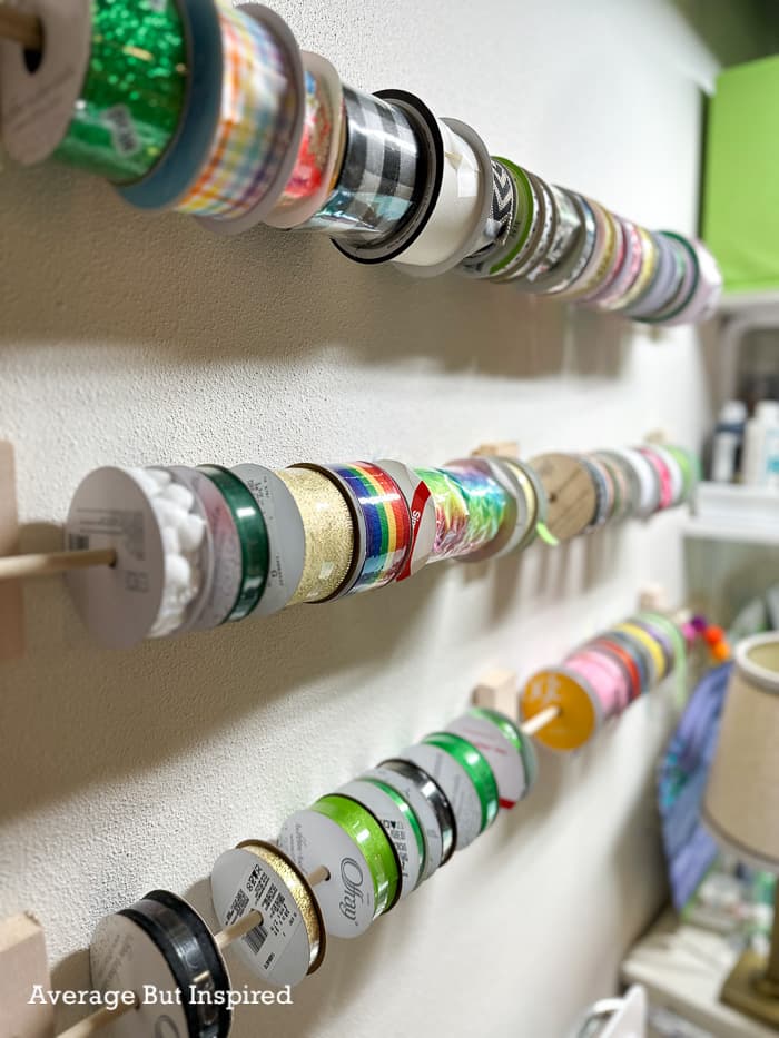 Rows of colorful ribbon spools on wooden dowels held by small painted blocks and eye hooks on a white craft room wall
