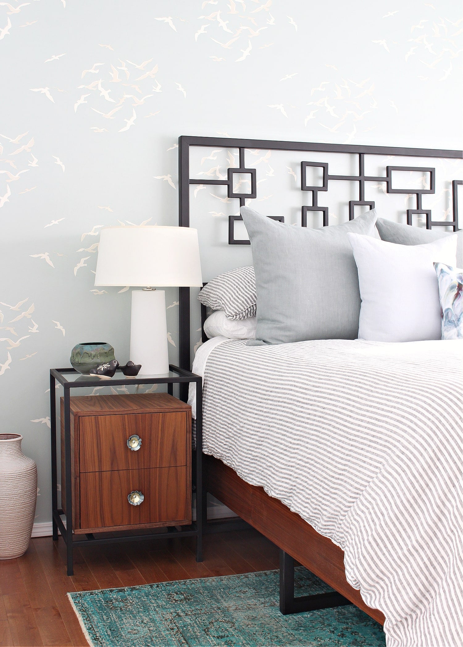 Walnut nightstand with two drawers in a black metal frame beside a bed with a geometric headboard and a white lamp