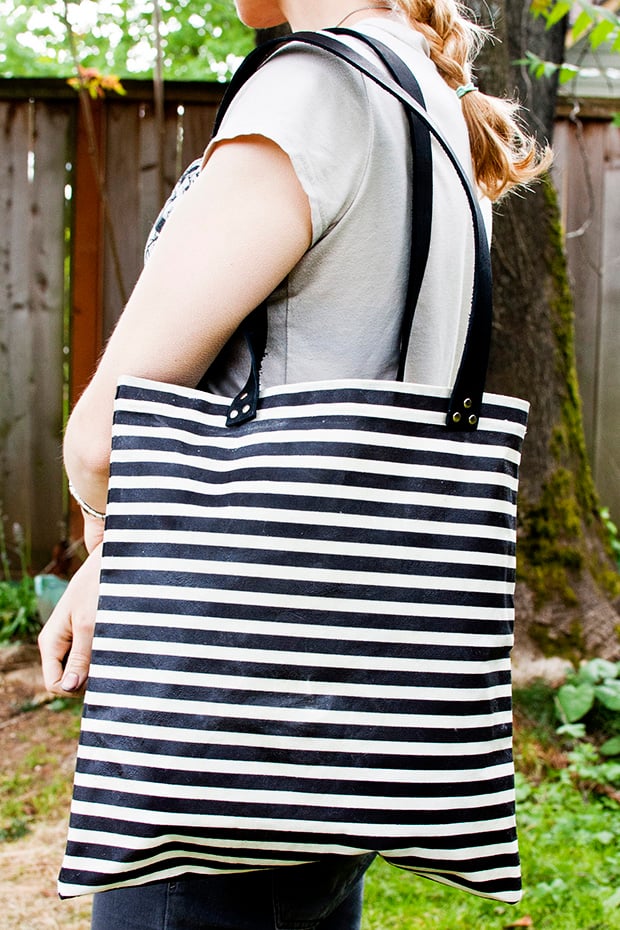 Black and white striped waxed canvas tote bag with black leather shoulder straps and rivets, carried on the shoulder outdoors