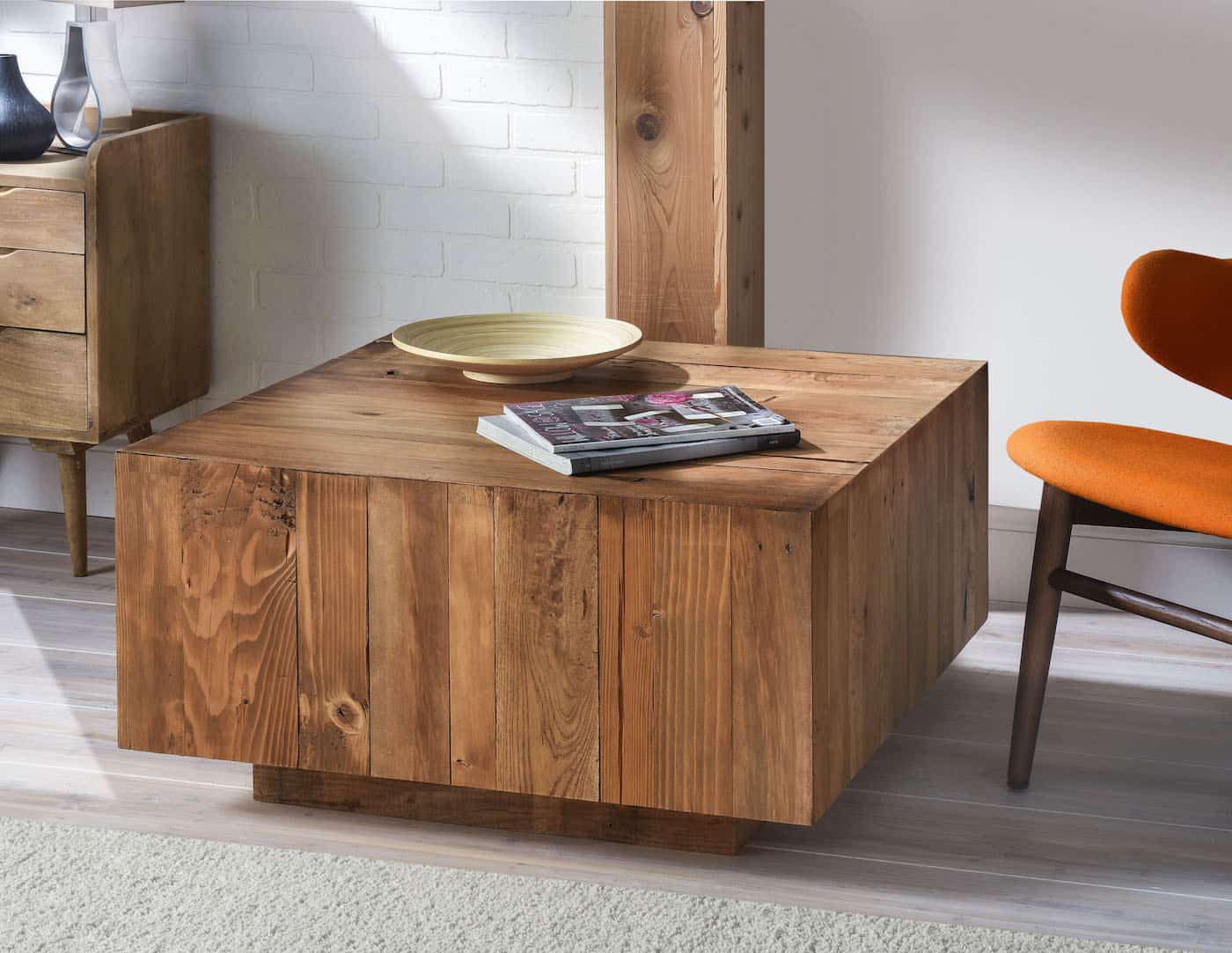 Square pallet wood plank coffee table with a warm stain, holding a plate and magazine beside an orange chair in a living room