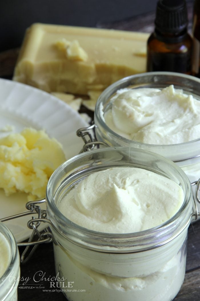 Glass jars of fluffy whipped white homemade body butter with chunks of raw shea butter set beside them on the table