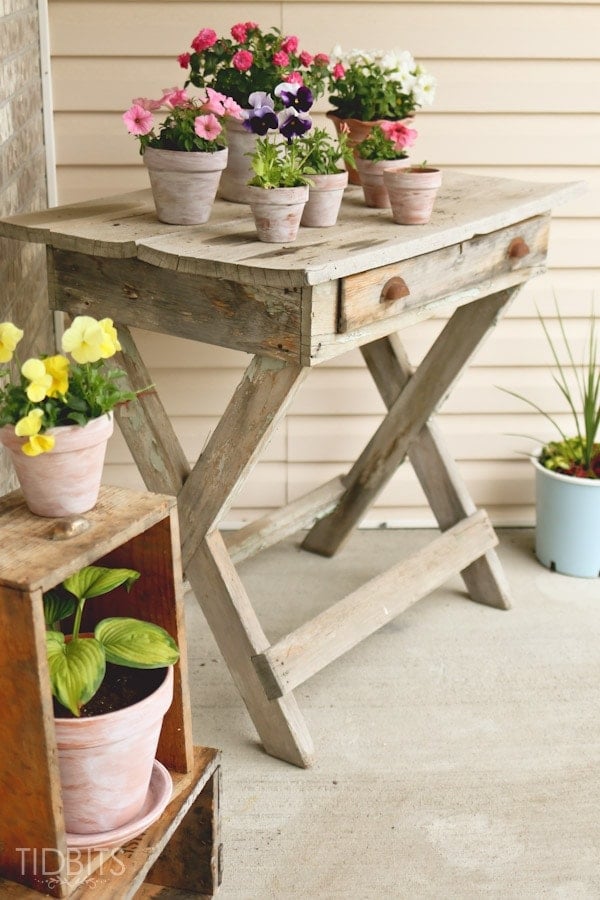 White washed terra cotta pots of pink, white and purple flowers on a weathered wooden X leg table on a front porch