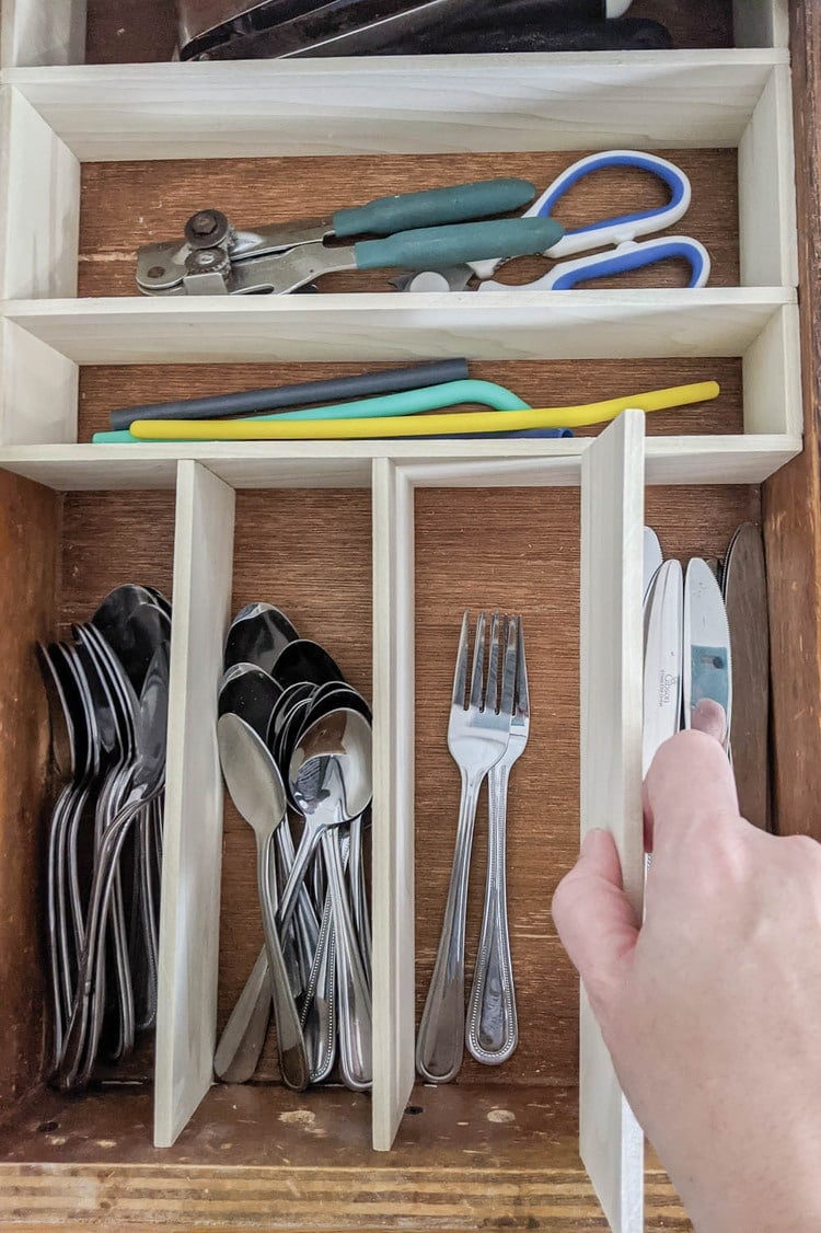 Kitchen drawer with thin wood dividers being fit in place around spoons, forks, knives, scissors and a can opener