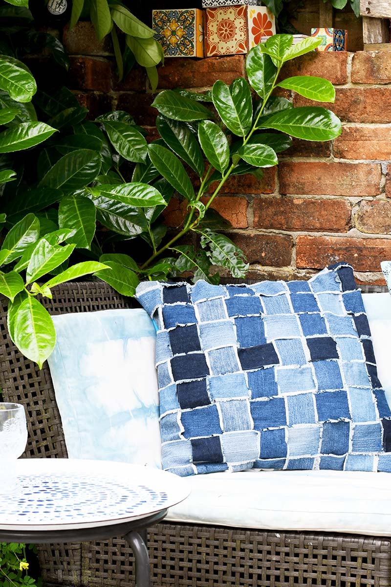 Woven denim throw pillow with frayed strips in light, medium and dark blue on a white cushion of a wicker garden chair