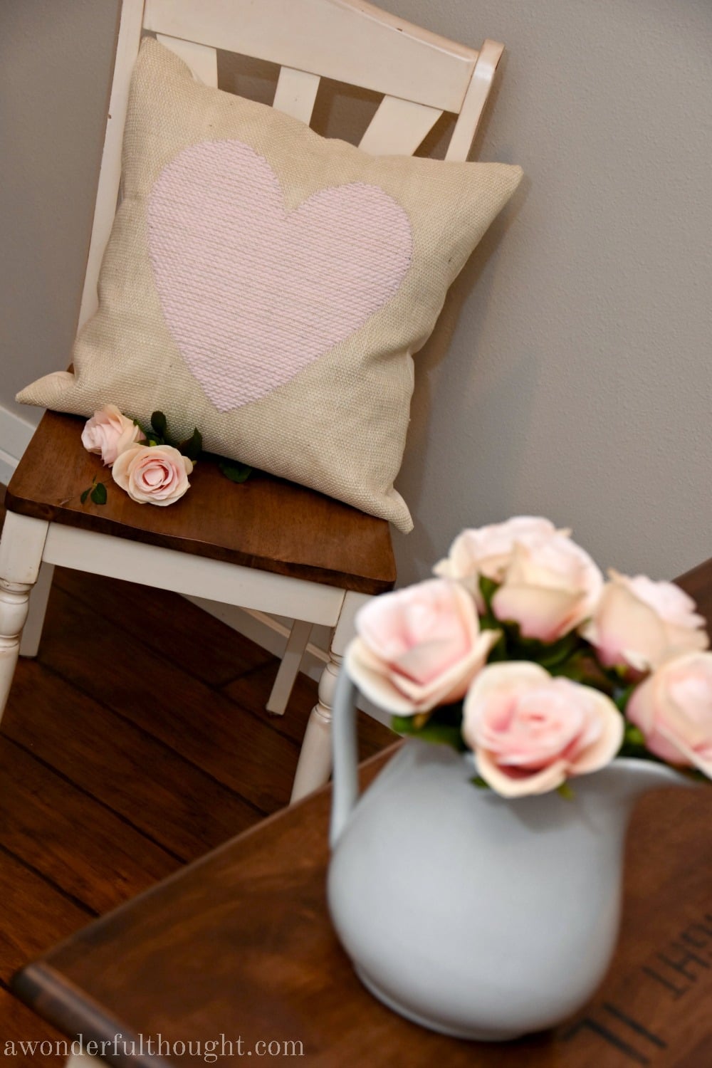 Cream burlap pillow with a pale pink yarn heart woven into the front on a wood chair beside a pitcher of pink roses